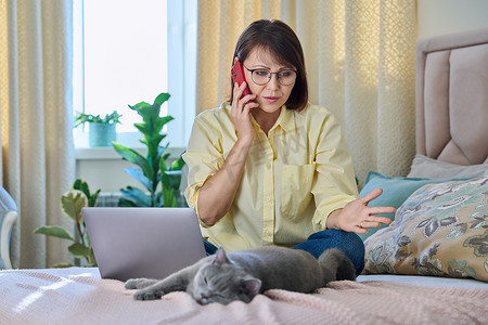 中年妇女坐在床上，带着笔记本电脑，猫在电话里聊天