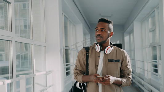 智能手机手机摄影照片_戴着白色大耳机的帅气非洲裔美国男学生拿着智能手机在大学明亮的长廊里散步，给某人发短信