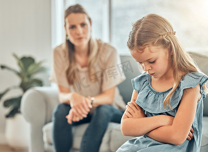 悲伤的白人小女孩坐在客厅里，双臂交叉，感到沮丧，而她的母亲则生气地看着。
