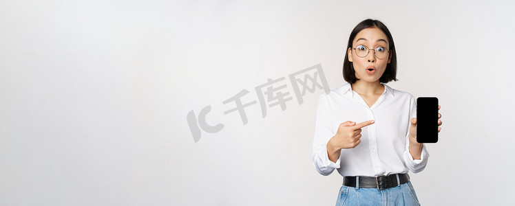 公司背景展示摄影照片_热情的亚洲年轻女性用手指指着智能手机屏幕，在手机上展示广告，白色背景