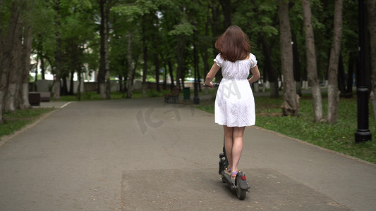 身穿白色连衣裙的年轻亚洲女性在公园里骑着共享电动滑板车。