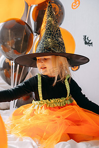 儿童万圣节-一个戴着女巫帽的女孩和一个狂欢节服装，家里有通风的橙色和黑色气球。