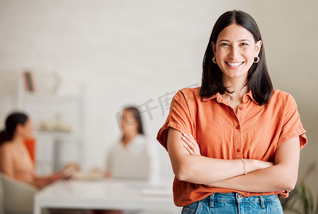 一位自信的西班牙裔年轻女商人双臂交叉站在办公室里，背景是她的同事。