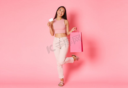 优惠客户摄影照片_无忧无虑、梦幻般可爱的亚洲女孩，在粉红色背景下摆着信用卡和购物袋，在购物中心享受周末，周围有特别优惠和折扣