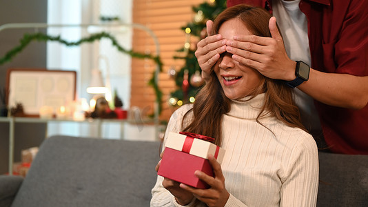 家庭欢庆摄影照片_惊讶的年轻女子坐在沙发上，手里拿着一个礼品盒，而她的男朋友站在后面，用手遮住眼睛