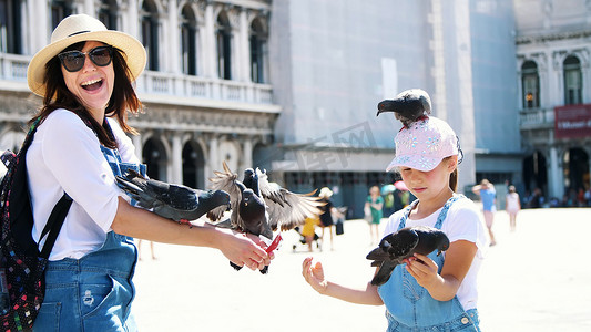 夏日玩乐摄影照片_意大利威尼斯-2018 年 7 月 7 日：夏日，快乐的女人和小女孩、游客、抱着鸽子、喂食、和它们玩耍、在圣马可广场、圣马克大教堂玩乐