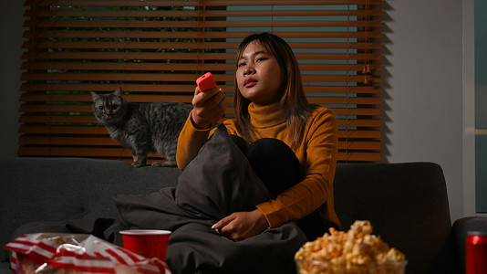 沙发猫摄影照片_带遥控器的年轻女士在舒适的客厅里和她的猫坐在沙发上欣赏有趣的电影