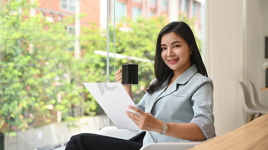 优雅的年轻职业女性在明亮的现代办公室喝咖啡和阅读文件