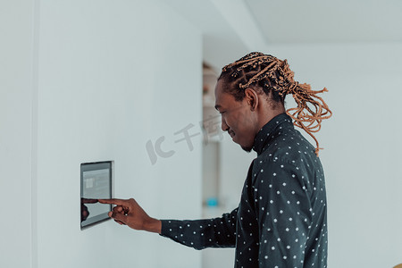 智能家居系统摄影照片_微笑的非洲裔美国人使用现代智能家居系统、墙上的控制器、积极的年轻人在恒温器上切换温度或激活公寓的安全警报