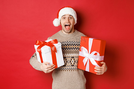 欢快迷人的男人拿着圣诞礼物，戴着圣诞帽和冬日毛衣，微笑着惊讶地站在红色背景上的形象