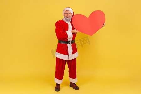身穿圣诞老人服装、手持大红心、留着灰胡子的老人的全长肖像，表情积极地看着相机。