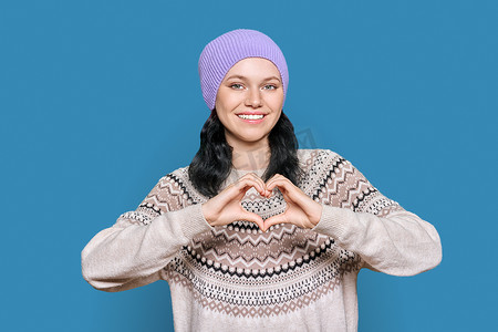 身穿帽子毛衣的年轻少女双手在蓝色背景上展示心脏姿势