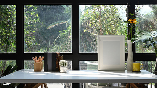 家庭办公室玻璃窗附近的白色桌子上的相框、咖啡杯、书籍和犁壶。