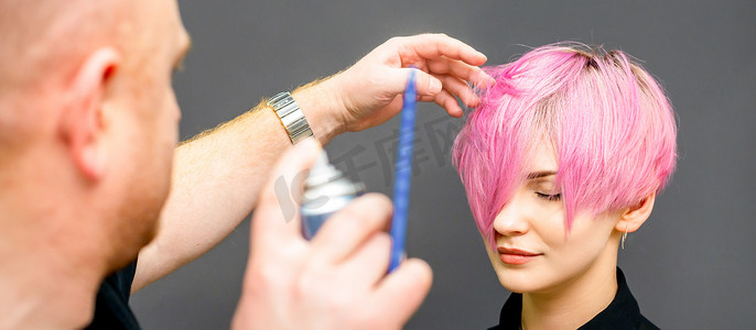 美发师正在使用发胶固定美发沙龙中年轻白人女性的粉红色短发。