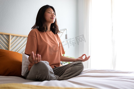 年轻的亚洲女性穿着舒适的衣服，早上在床上冥想以清醒头脑。