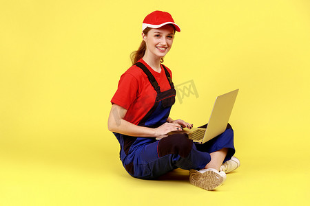 满意的得心应手的女人盘腿坐在地板上，在笔记本电脑上工作，看着相机。
