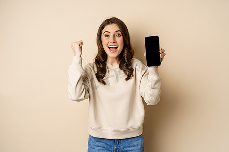 热情的女性欢呼并展示手机屏幕、智能手机应用程序界面，站在米色背景上
