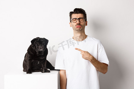 悲伤摄影照片_悲伤而阴郁的宠物主人指着他的黑色哈巴狗抽泣，站在白色背景下