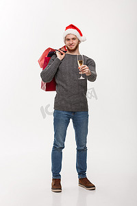 冬天购物袋摄影照片_圣诞节概念-年轻英俊的胡子男子拿着一杯香槟和购物袋，脸上表情快乐。