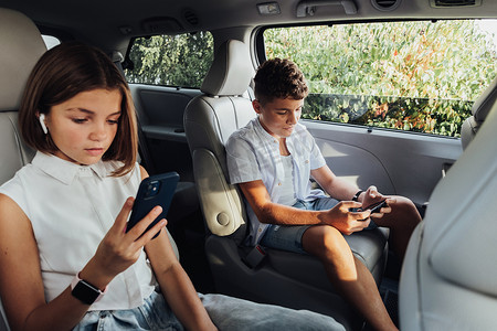 年轻的白人男孩和他十几岁的妹妹坐在小货车里专注于智能手机，孩子们在周末家庭自驾公路旅行中玩游戏