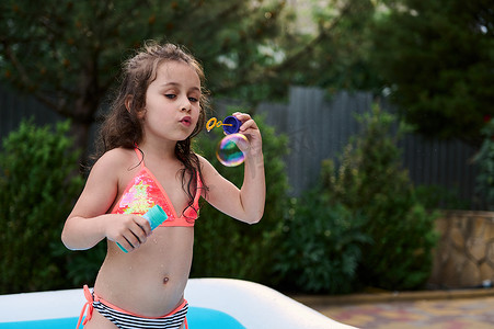 穿着时髦泳衣的可爱 5 岁小女孩在家庭花园的充气儿童泳池里吹肥皂泡