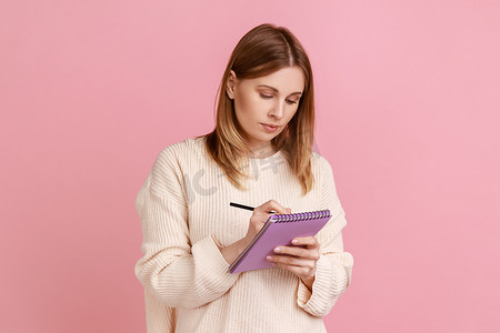 集中精力的金发女性在纸质笔记本上写字，制作待办事项清单或购买清单。