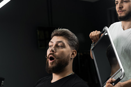 剃胡子的男人摄影照片_快乐而惊讶的留着胡子的男人在理发店照镜子。