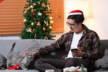 沙发猫摄影照片_戴着圣诞老人帽子的年轻人和他的猫坐在家里的沙发上。