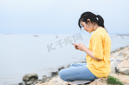 摘口罩摄影照片_年轻的亚洲女性在接种 Covid-19 或冠状病毒疫苗后，正在海边使用智能手机，摘下口罩呼吸新鲜空气。