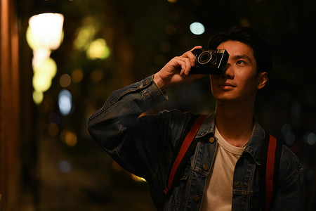 时髦的男子旅行者站在夜间街灯背景模糊的城市里用复古相机拍照