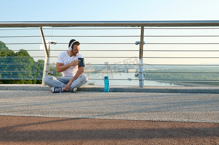 电子健康卡摄影照片_年轻运动员、戴耳机的运动员坐在城市桥梁上，检查移动应用程序，在智能手机支架上用手机锻炼时监测心率和燃烧的卡路里