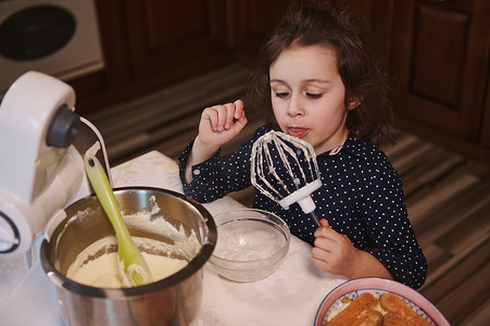 甜甜摄影照片_可爱的白人小女孩在她母亲不在家庭厨房时，从食品加工机搅拌机中舔着甜甜可口的生奶油