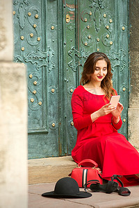 美丽的年轻白人女子坐在门口的楼梯上，带着旅行箱和智能手机，穿着红色长裙。