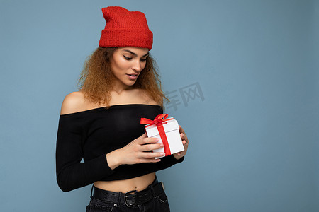 照片中，漂亮迷人的年轻卷发深色金发女郎孤立地站在蓝色背景墙上，上面有文字复制空间，穿着日常时尚的黑色上衣和红色帽子。