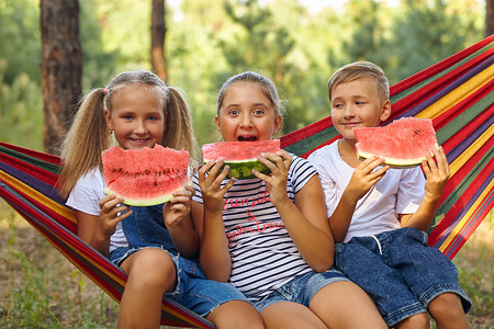 开玩笑的摄影照片_孩子们坐在吊床上吃西瓜，在新鲜空气中开玩笑
