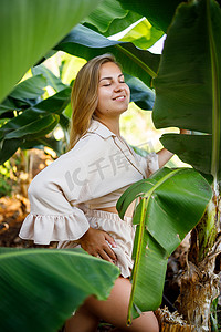 在热带地区的一个公园里，靠近香蕉灌木绿叶的女人，她穿着米色裙子和衬衫