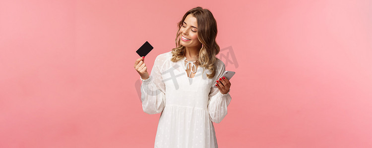 身穿白裙的快乐漂亮年轻金发女性的肖像，对信用卡看起来很高兴和满意，手持智能手机，推荐在线购买，互联网购物概念