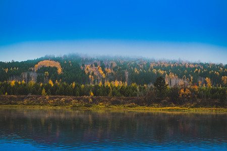 秋天河岸的景观，山上覆盖着茂密的绿叶和黄叶森林。