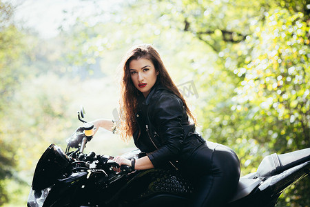 在阳光明媚的天气里，在大自然、街道、森林里，一位穿着黑色皮衣骑着摩托车的漂亮年轻女子的肖像，特写，看着相机