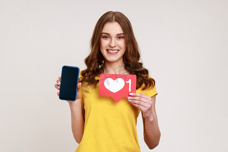 主图标摄影照片_积极微笑的少女博主拿着类似的图标，展示带有空白屏幕的智能手机，在社交网络上发帖并等待反馈。