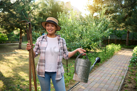 充满灵感的女园艺师手里拿着喷壶、锄头和裂土器，在春日在有机农场享受园艺