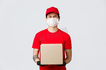 厌恶摄影照片_愤怒的亚洲送货员戴着红帽、T恤、手提箱，戴着医用口罩和手套，抱怨粗鲁的客户，皱着眉头厌恶和厌恶。