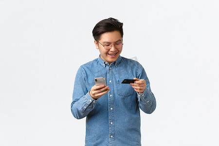 戴着眼镜、穿着休闲服、微笑着、无忧无虑的亚洲男人，戴着牙套，高兴地看着信用卡，用智能手机应用程序在网上购物，用手机在网上购物