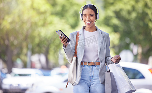 营销技巧摄影照片_在城市购物，戴着智能手机和耳机的女性面带微笑地收听时尚服装播客，了解营销销售技巧和风格理念。