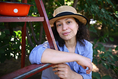 一个宁静美丽的女人的肖像，戴着草帽的农民，靠在樱桃园的梯子上，看着相机