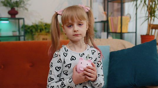 悲伤的贫穷小女孩摄影照片_沮丧的可怜小女孩拿着几乎空的存钱罐，只有一美元现金，破产