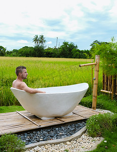 寄宿摄影照片_泰国一家寄宿家庭里的男人在外面的浴缸里度假，那里有绿色的稻田