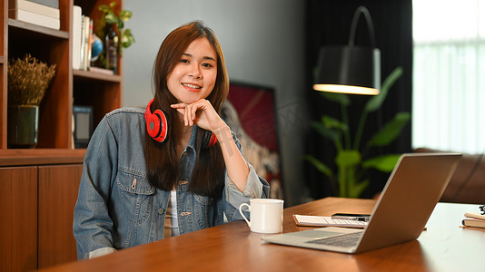 坐着的会计摄影照片_戴着耳机的迷人年轻女性坐在舒适的家庭办公室里，对着镜头微笑