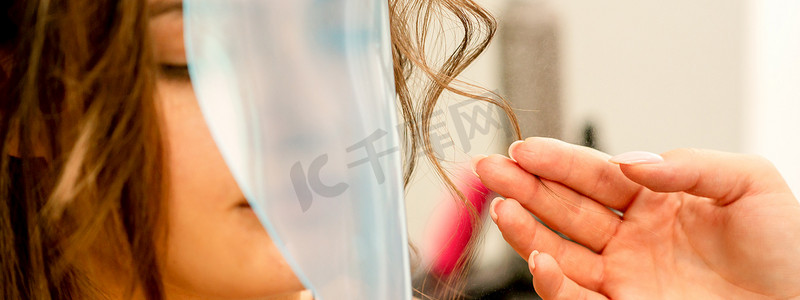 专业美发师在美容院用发胶造型和固定长发女性卷发，用防护罩覆盖脸部。