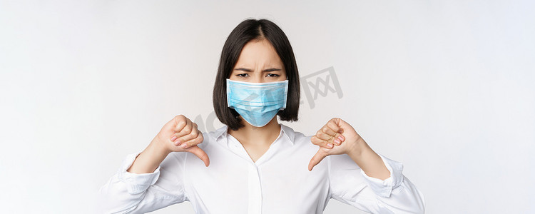 疲惫背景摄影照片_戴着医用面罩的亚洲女性的肖像，站在白色背景上，大拇指朝下，表情失望、疲惫